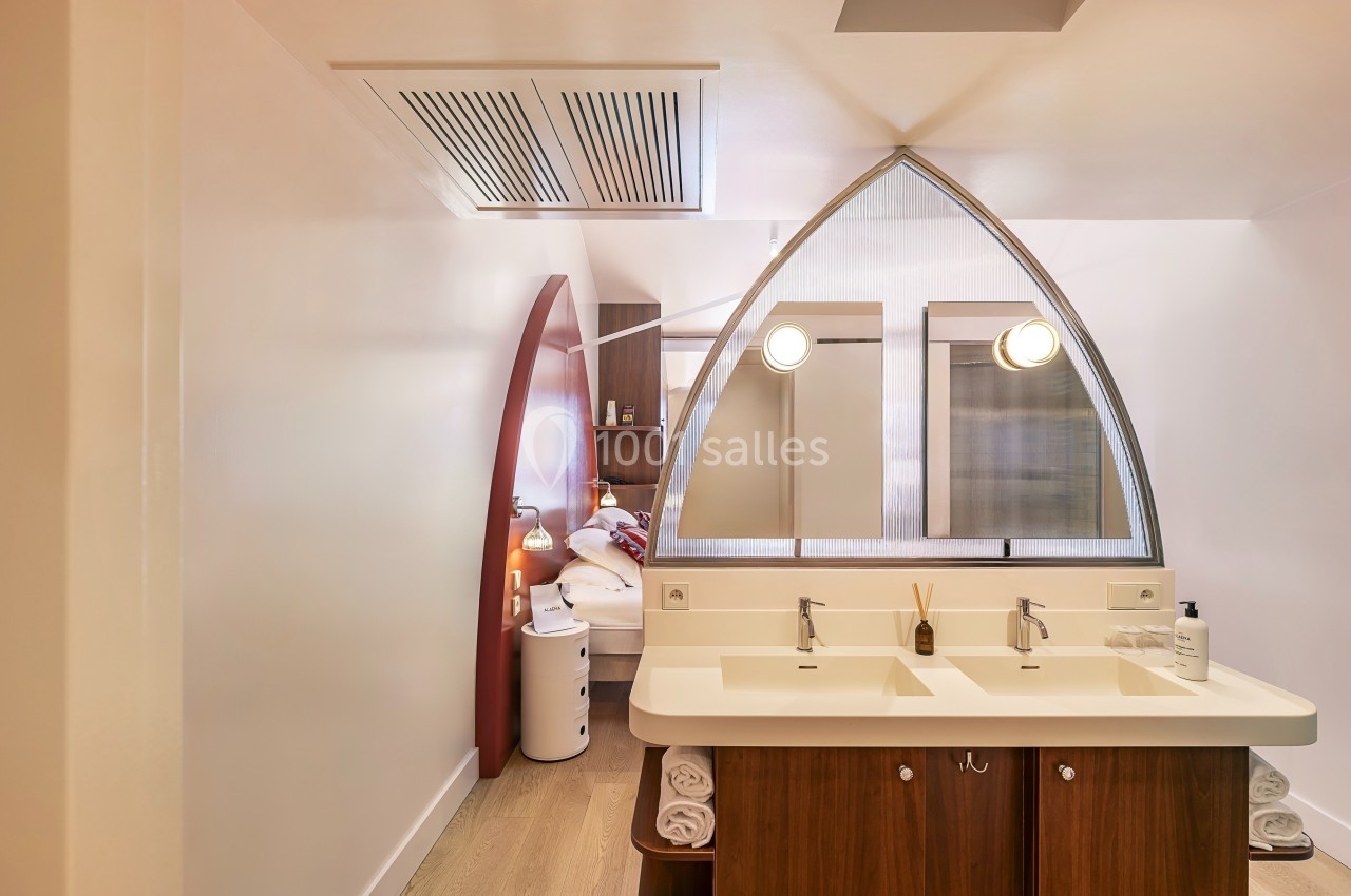 Salle de bain moderne avec double vasque, miroir en arche et vue partielle sur une chambre à travers une cloison vitrée.