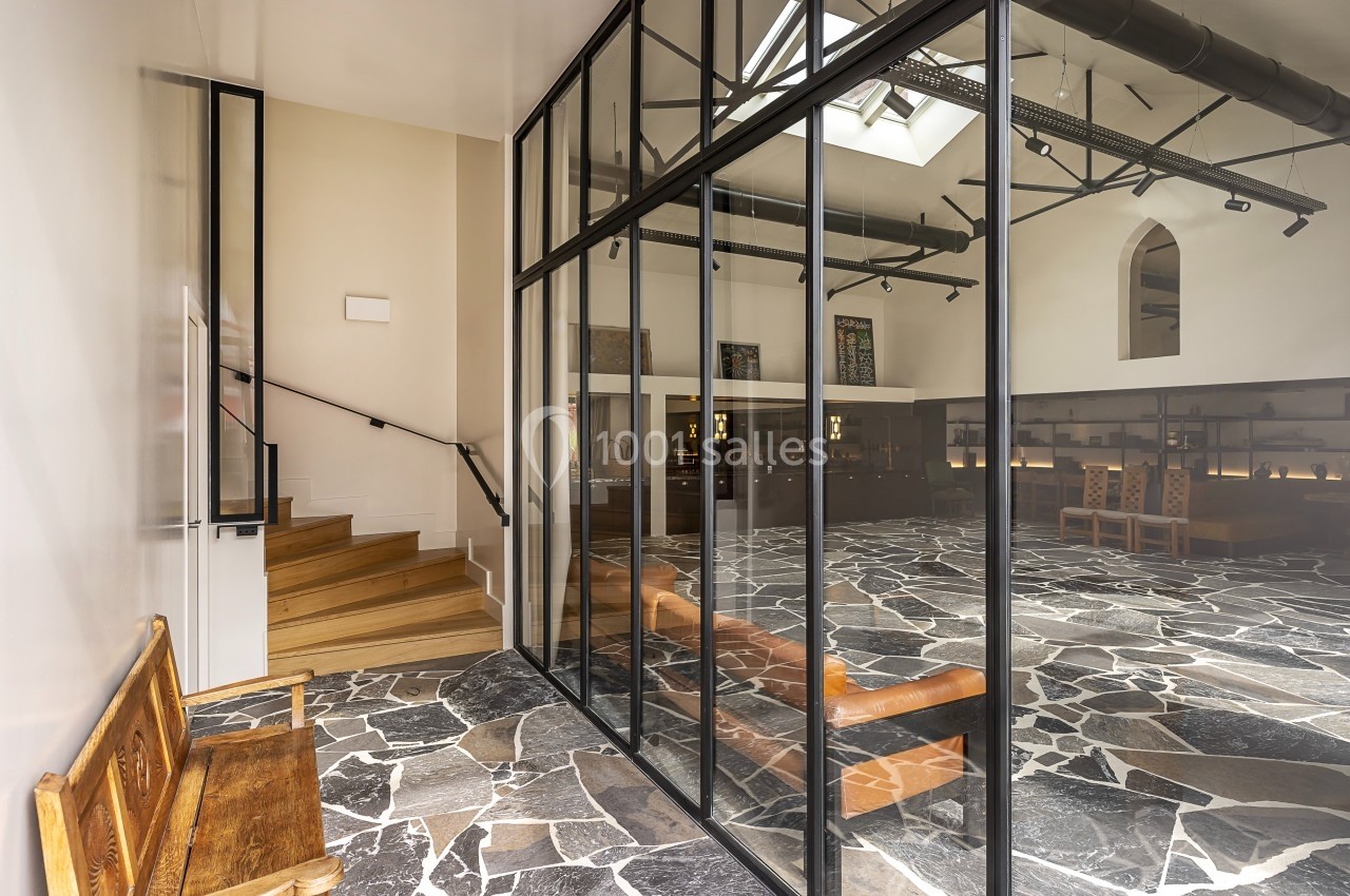 Escalier en bois et cloison vitrée donnant sur une pièce avec sol en pierre et mobilier en bois.