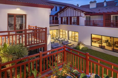 Vue d'une cour intérieure avec balcons en bois rouge, grandes baies vitrées et éclairage chaleureux au crépuscule.