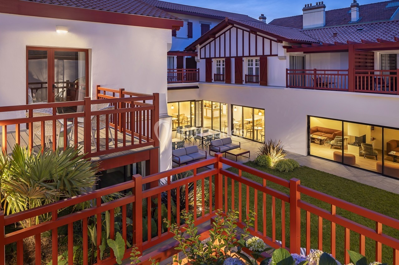 Vue d'une cour intérieure avec balcons en bois rouge, grandes baies vitrées et éclairage chaleureux au crépuscule.