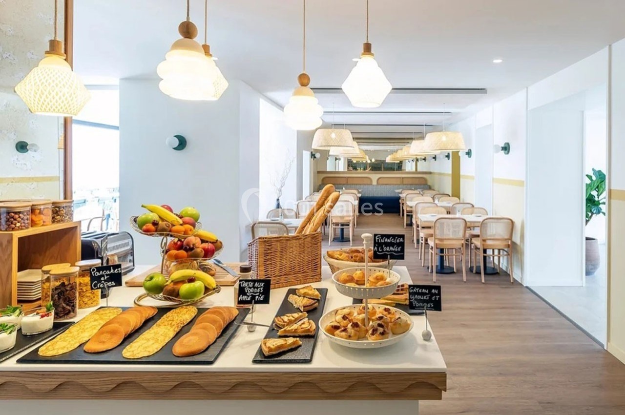 Buffet de petit-déjeuner avec viennoiseries, fruits, pains et céréales dans une salle lumineuse et moderne.
