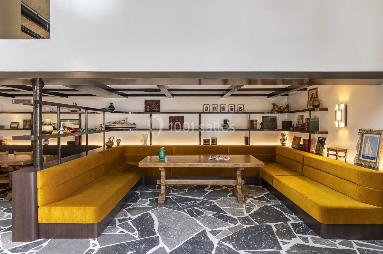 Salle avec banquettes jaunes en angle, table en bois au centre et étagères décorées d'objets et livres en arrière-plan.