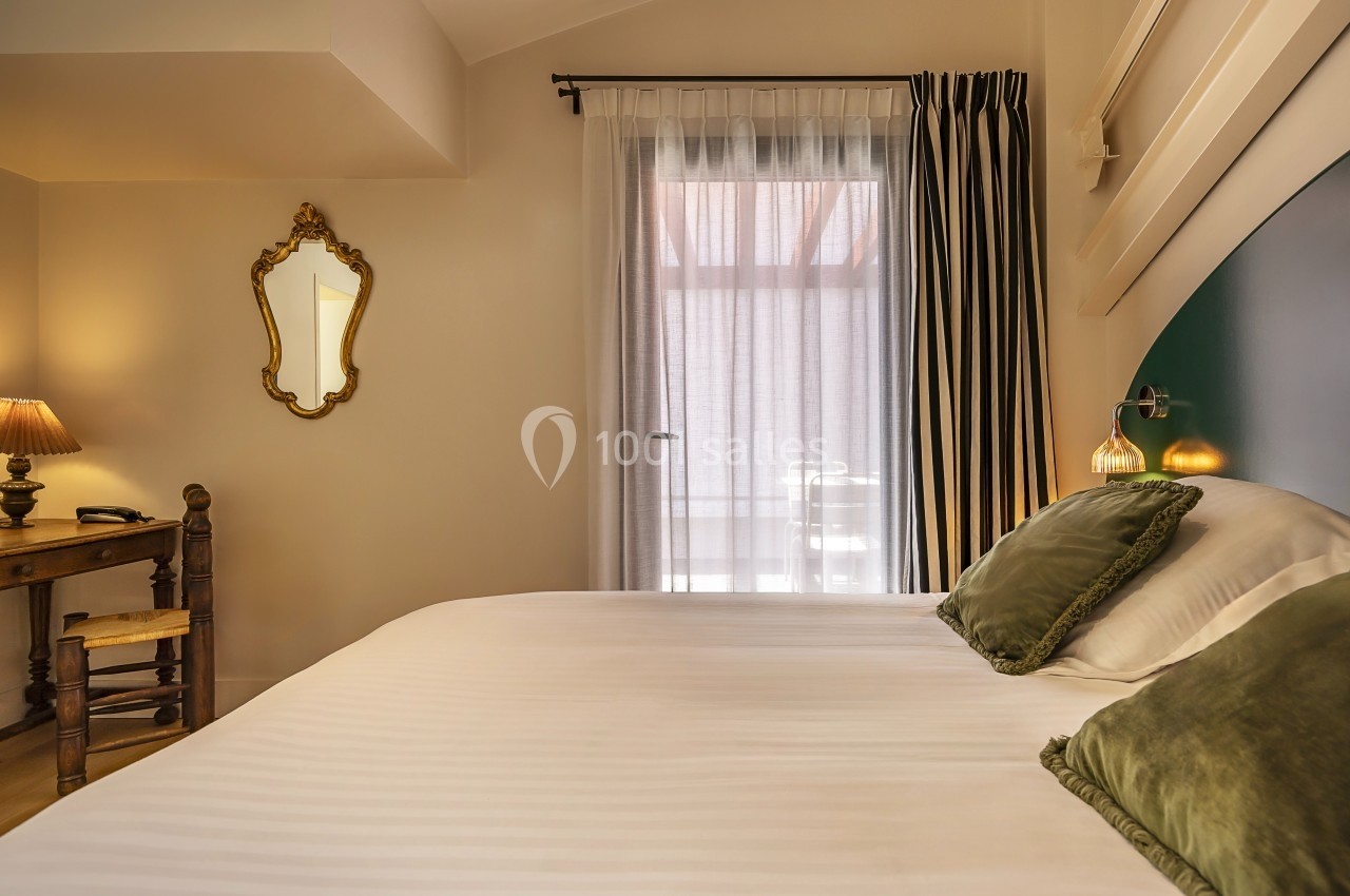 Chambre lumineuse avec lit double, coussins verts, bureau en bois, miroir doré et rideaux encadrant une porte vitrée.