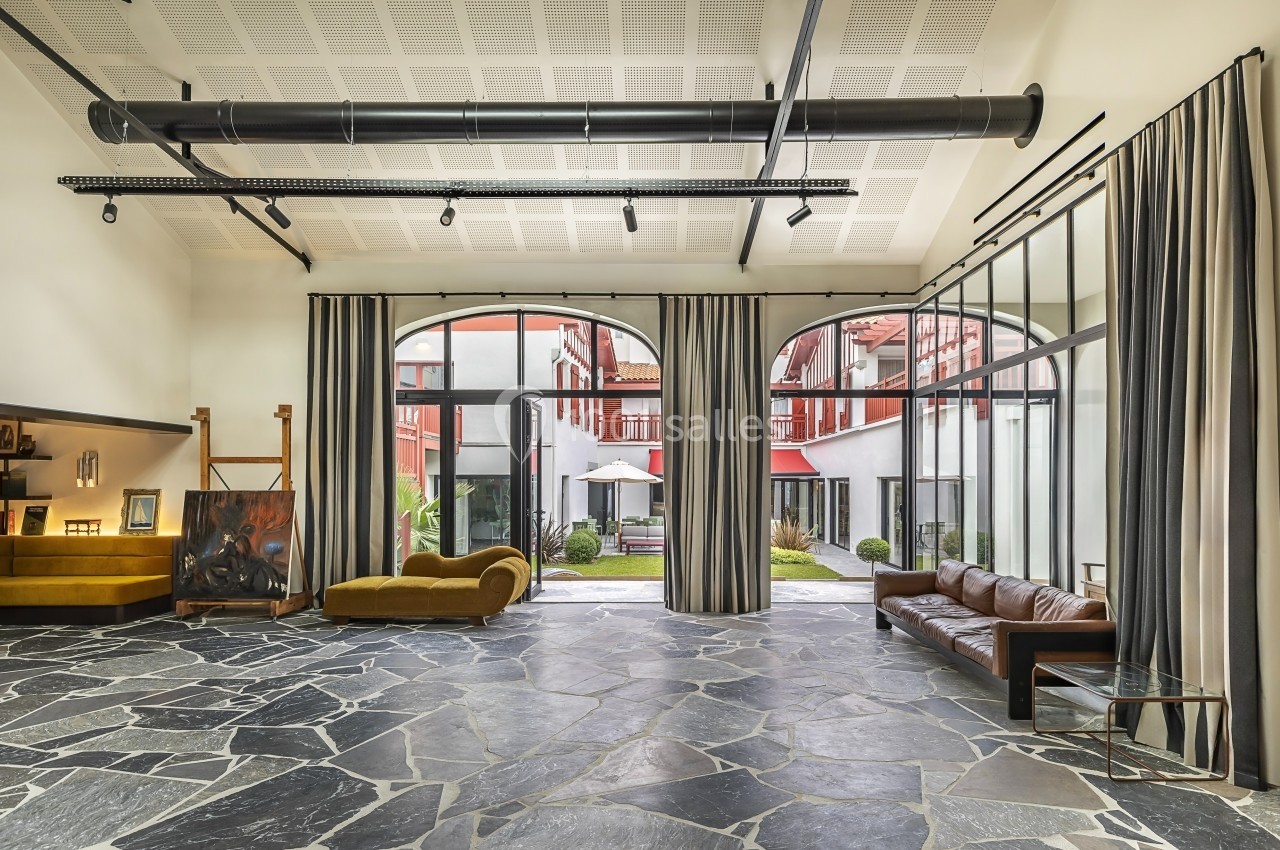 Salle lumineuse avec sol en pierre, canapés, grandes baies vitrées donnant sur une cour avec parasols et végétation.