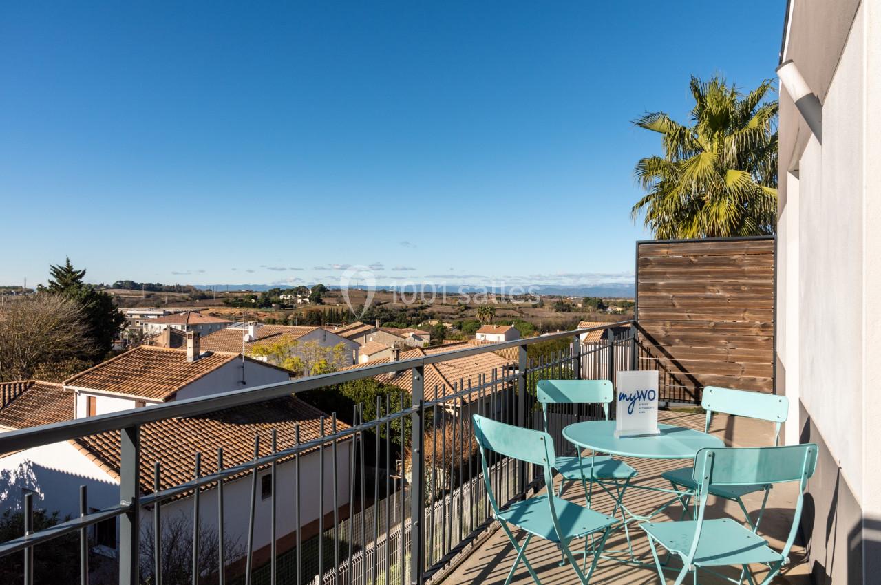 Vue depuis un balcon avec table et chaises bleues, donnant sur des toits de maisons et un paysage dégagé sous un ciel bleu.