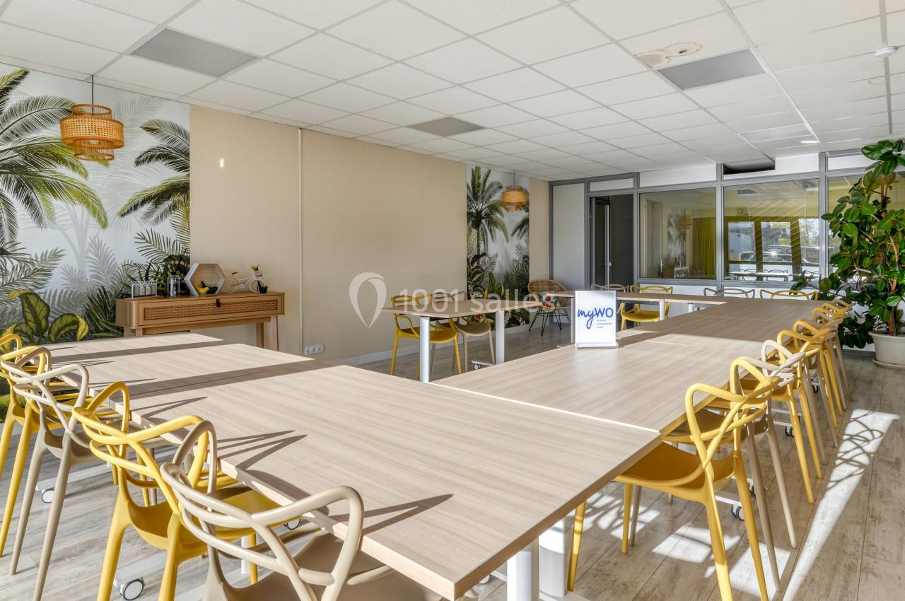 Salle de réunion lumineuse avec tables en U, chaises jaunes, plantes et décor mural tropical.