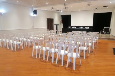 Salle de conférence avec des rangées de chaises blanches en plastique, un podium et un écran de projection.