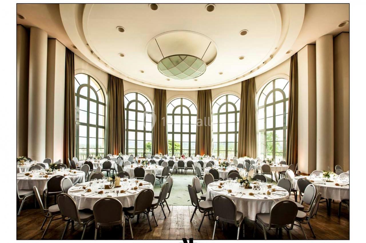 Salle de réception lumineuse avec de grandes fenêtres, des tables rondes dressées et des chaises grises.