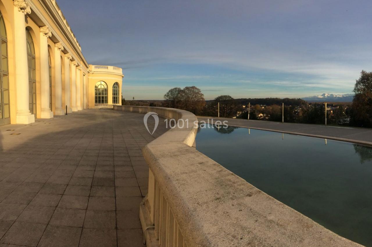 Terrasse avec balustrade longeant un bâtiment à colonnes, vue sur un bassin et un paysage lointain sous un ciel dégagé.