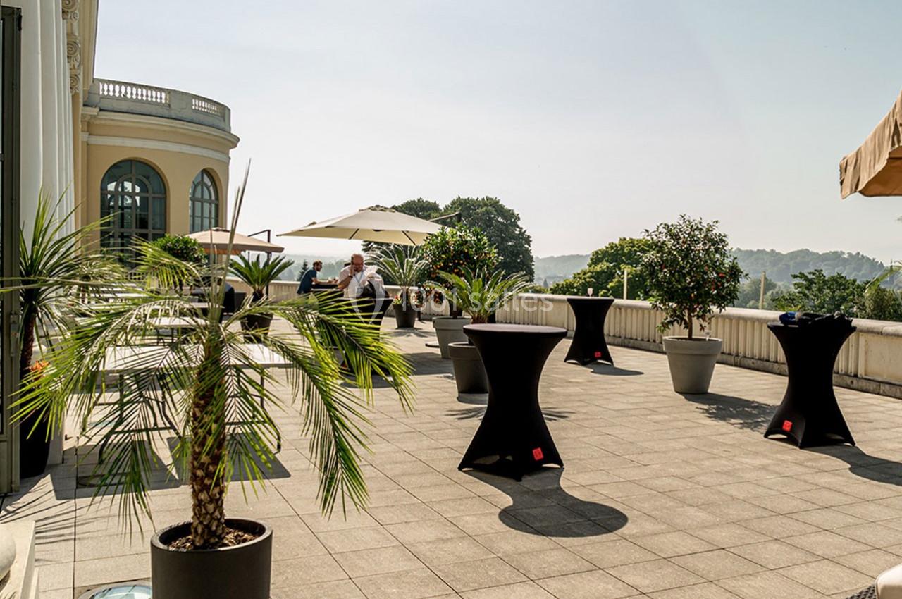 Terrasse ensoleillée avec tables hautes noires, plantes en pots et parasols, vue sur un paysage verdoyant.
