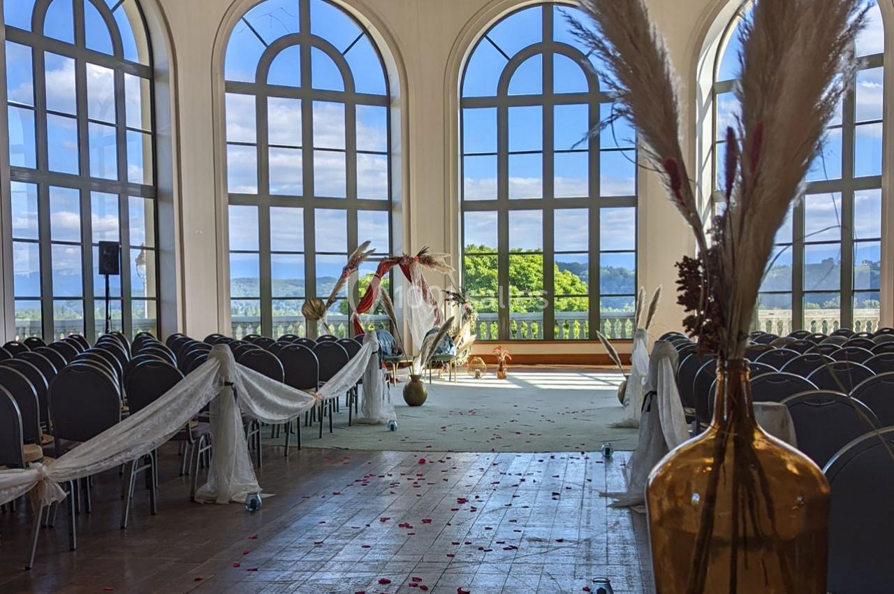 Salle de cérémonie lumineuse avec grandes fenêtres, rangées de chaises décorées et arche florale en arrière-plan.