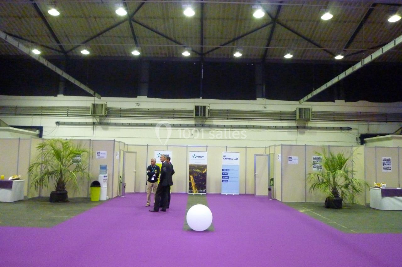 Trois personnes discutent au centre d'un espace d'exposition avec sol violet, stands en arrière-plan et éclairage suspendu.