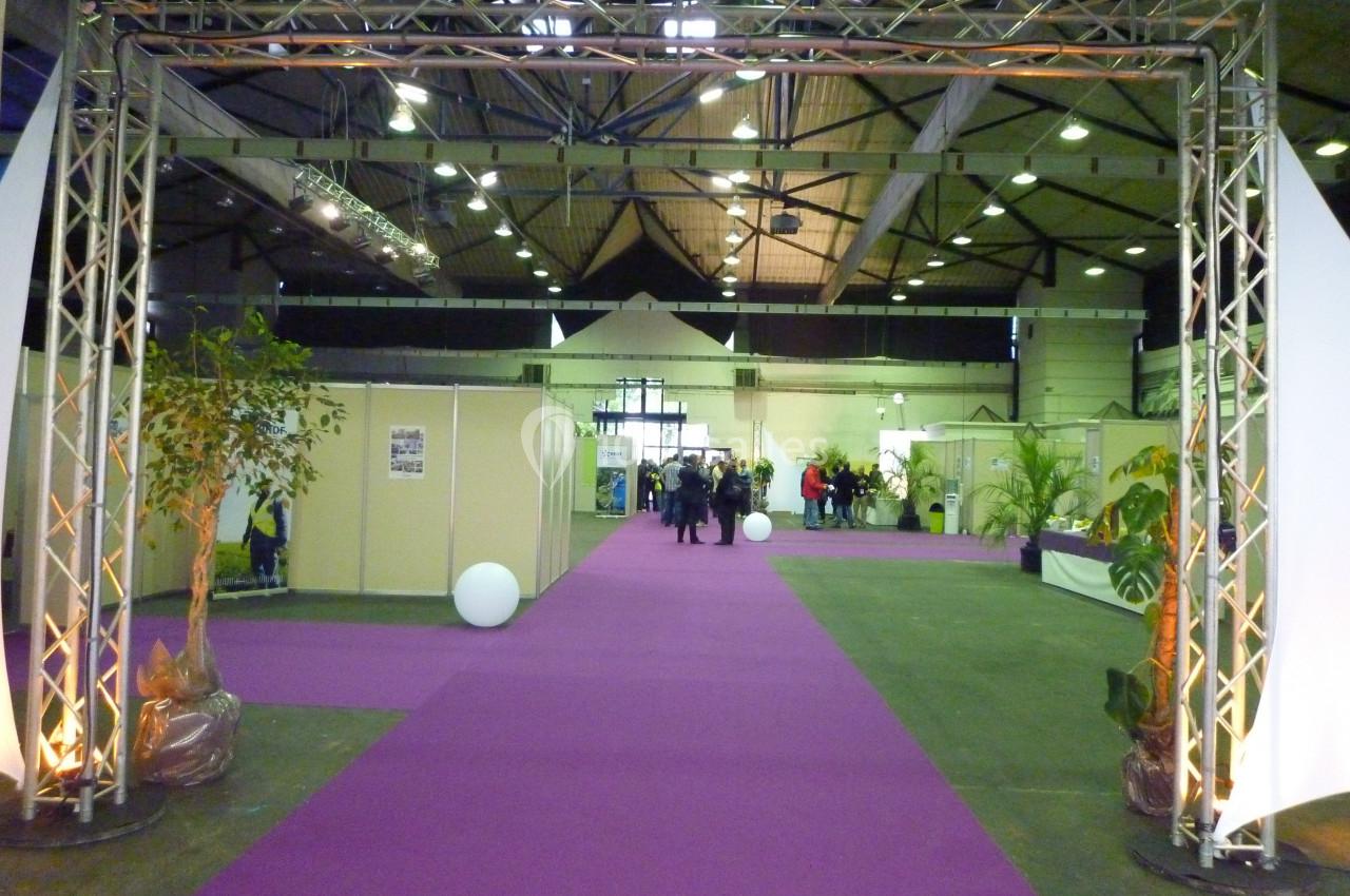 Allée centrale d'un salon ou d'une exposition avec moquette violette, structures métalliques et stands en arrière-plan.