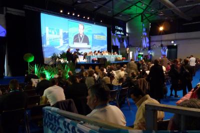 Salle de conférence avec des rangées de chaises blanches en plastique, un podium et un écran de projection.
