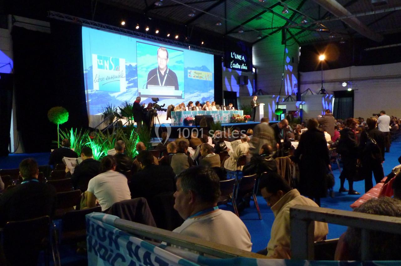 Salle de conférence avec un public assis, une scène éclairée et un écran projetant un intervenant.