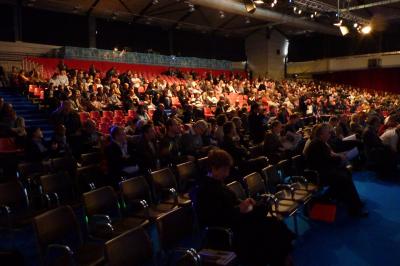 Salle de conférence avec des rangées de chaises blanches en plastique, un podium et un écran de projection.
