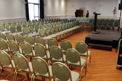 Salle de conférence avec des rangées de chaises blanches en plastique, un podium et un écran de projection.