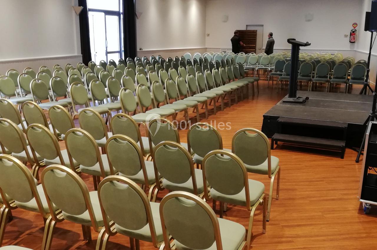 Salle de conférence avec rangées de chaises vertes alignées, une estrade avec pupitre et un équipement audio visible.