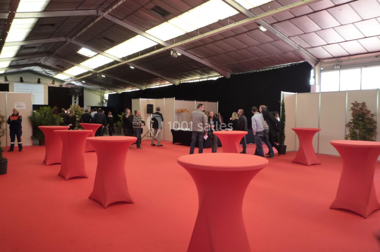 Salle d'exposition avec des tables hautes rouges et des visiteurs circulant près de stands.
