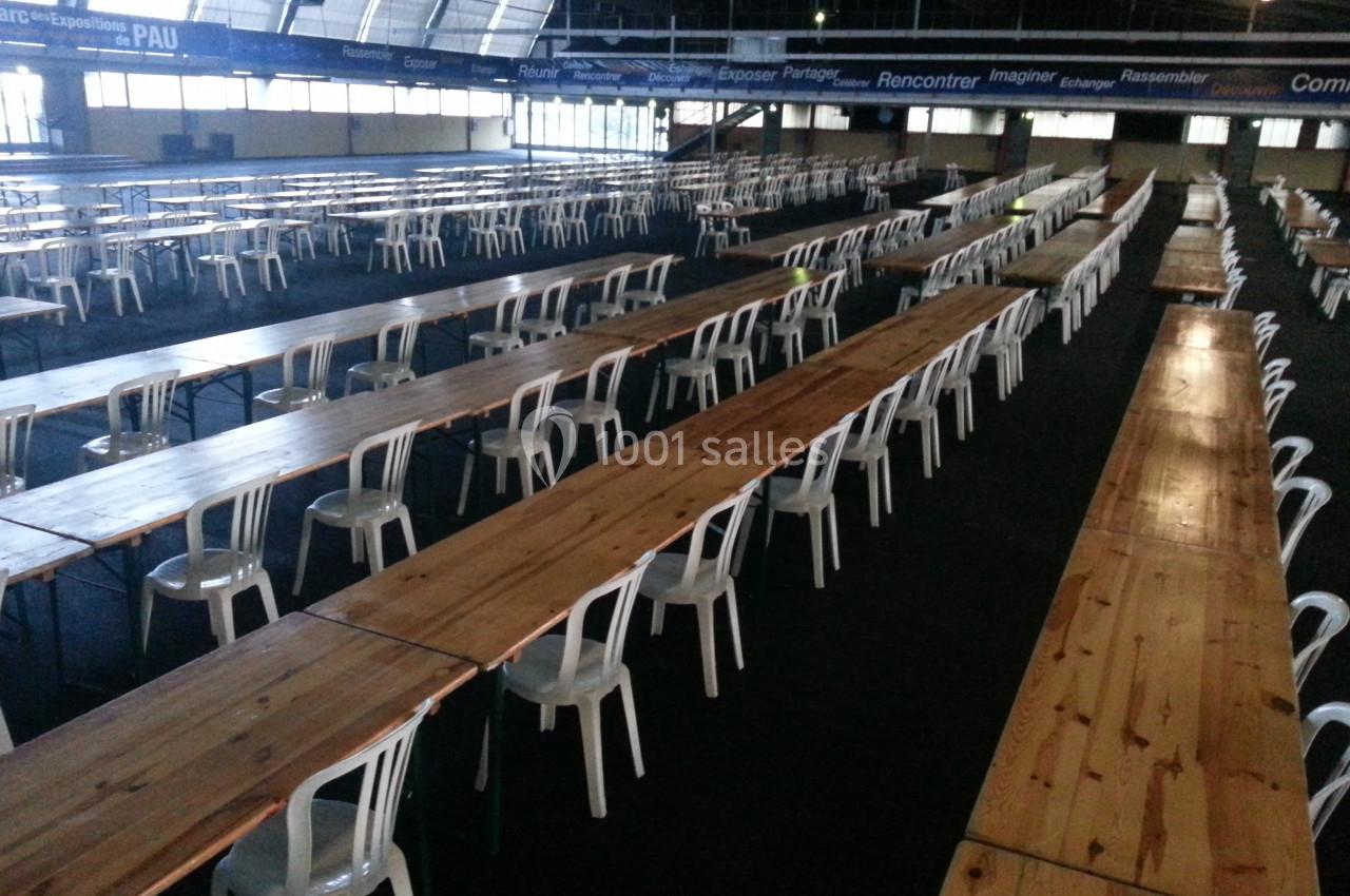 Salle avec de longues tables en bois alignées et des chaises blanches, prête pour un événement ou une réunion.