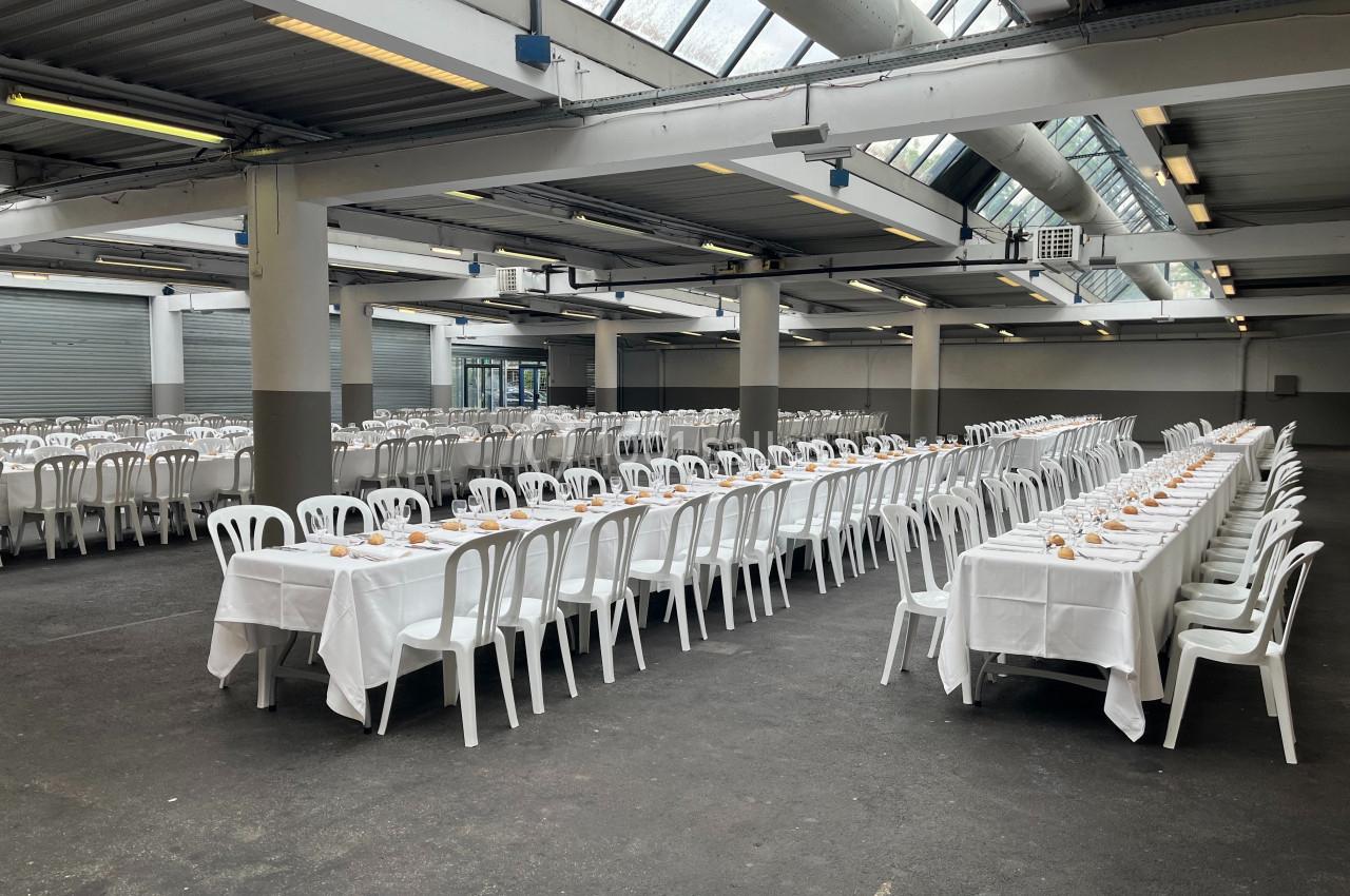 Grande salle avec de longues tables dressées de nappes blanches et alignées, entourées de chaises blanches en plastique.