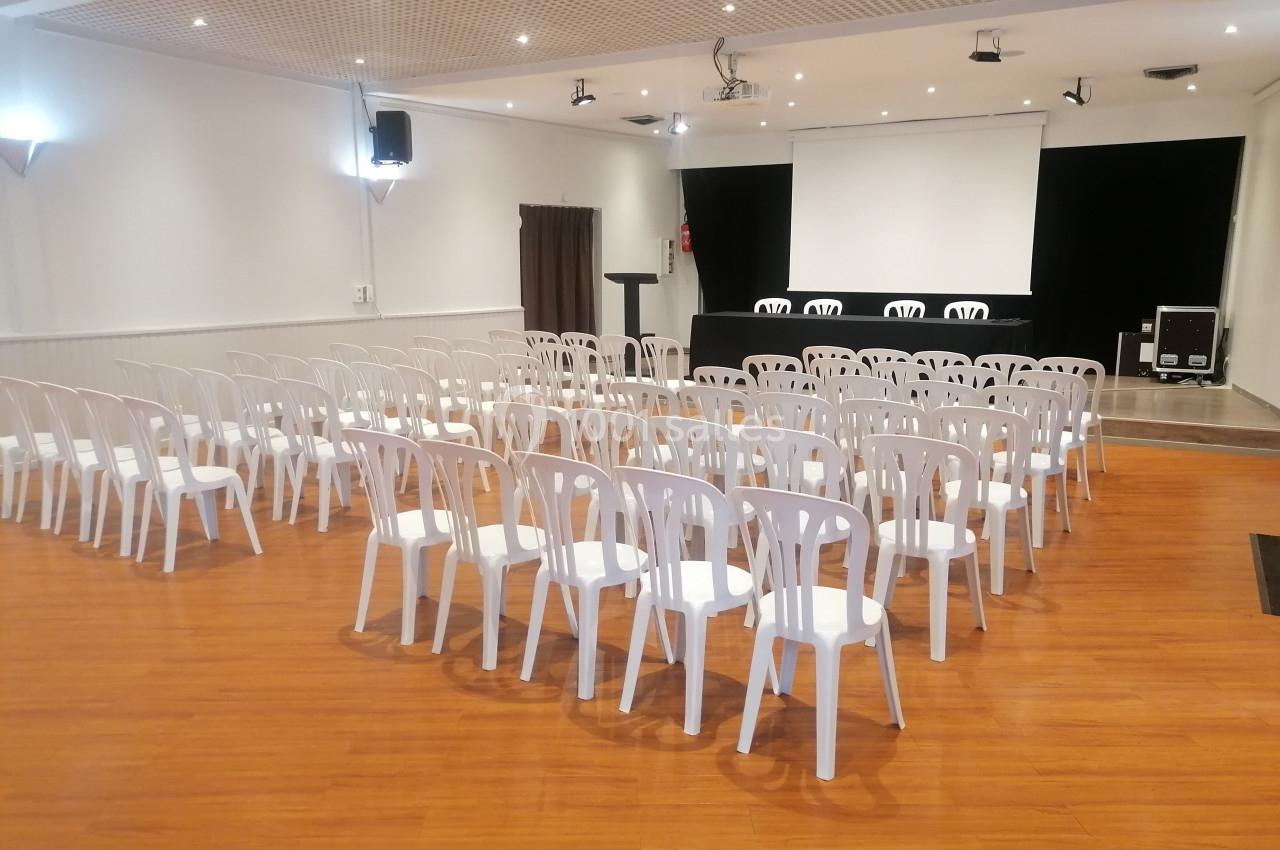 Salle de conférence avec des rangées de chaises blanches en plastique, un podium et un écran de projection.