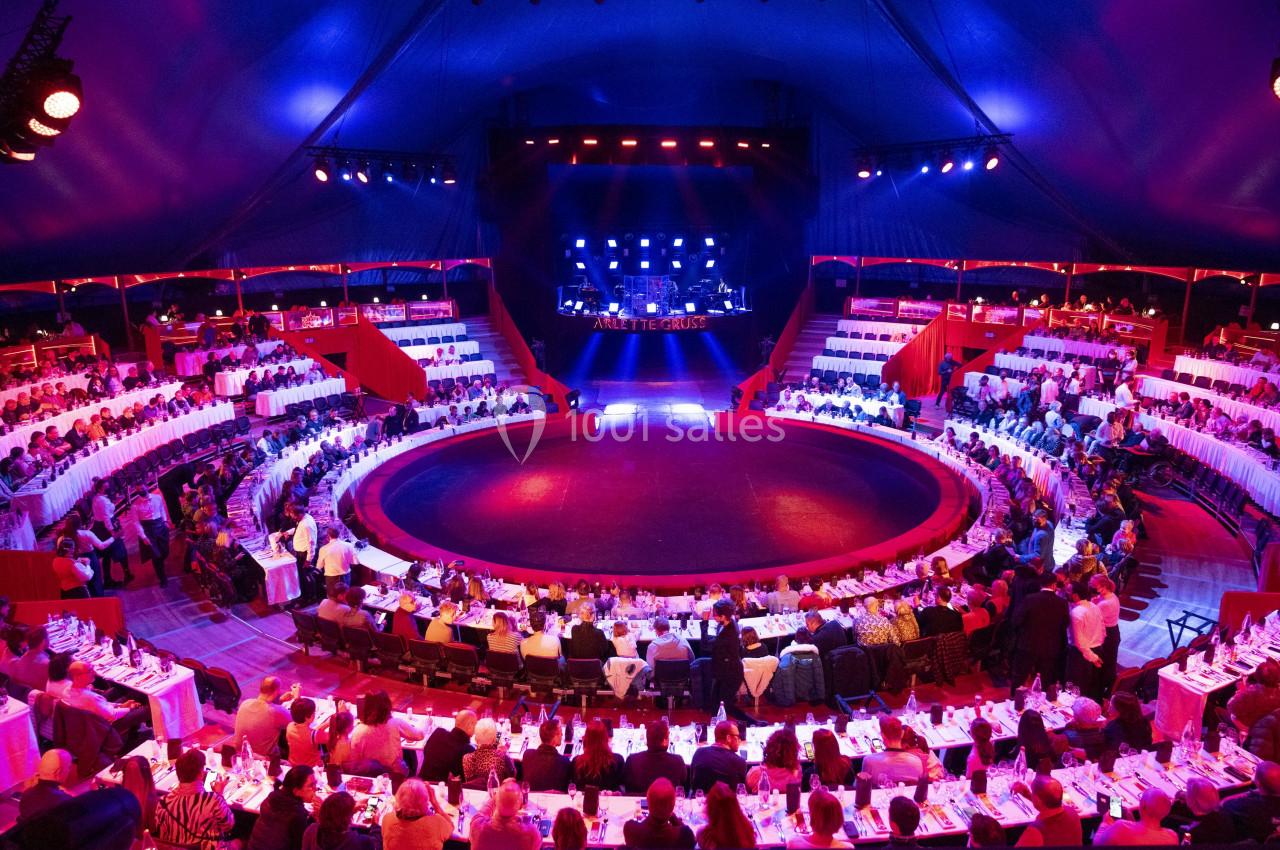 Vue intérieure d'un chapiteau de cirque avec des tables dressées autour d'une piste circulaire et une scène éclairée.