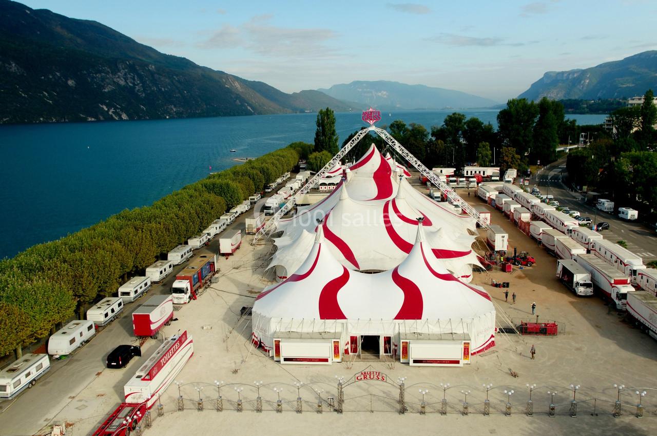 Vue aérienne d'un grand chapiteau blanc et rouge entouré de caravanes, près d'un lac et de montagnes.