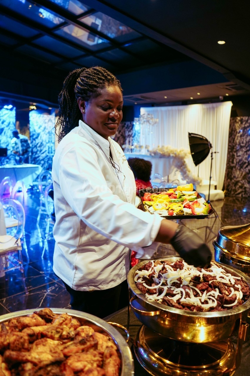 Une cheffe en tenue blanche dispose des plats garnis de viande et de légumes dans un buffet lors d'un événement.