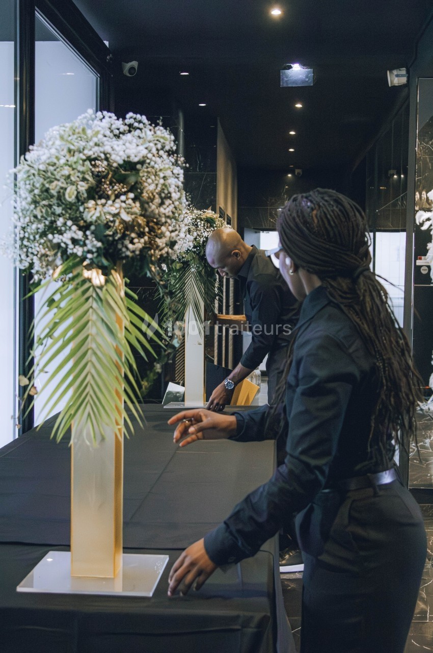 Deux personnes habillées en noir arrangent une table décorée avec un grand bouquet floral dans un espace intérieur sombre.