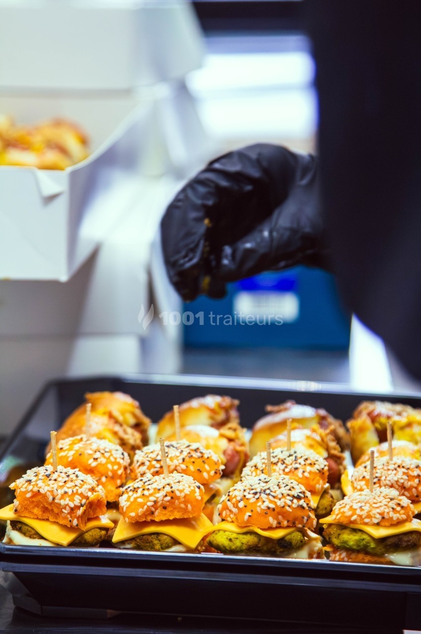 Plateau de mini-burgers garnis de fromage, sauce et graines de sésame, préparés par une main gantée.