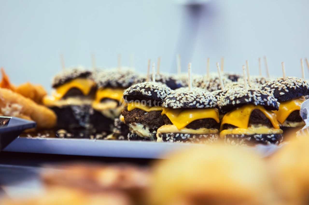 Mini-burgers au pain noir garnis de fromage fondu et de graines de sésame, alignés sur un plateau.