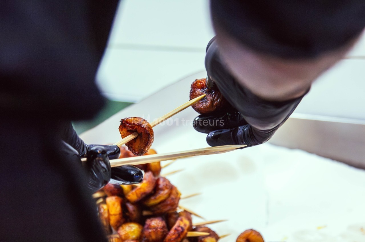 Personne en gants noirs en train de piquer des morceaux de bananes plantains grillées sur des brochettes.