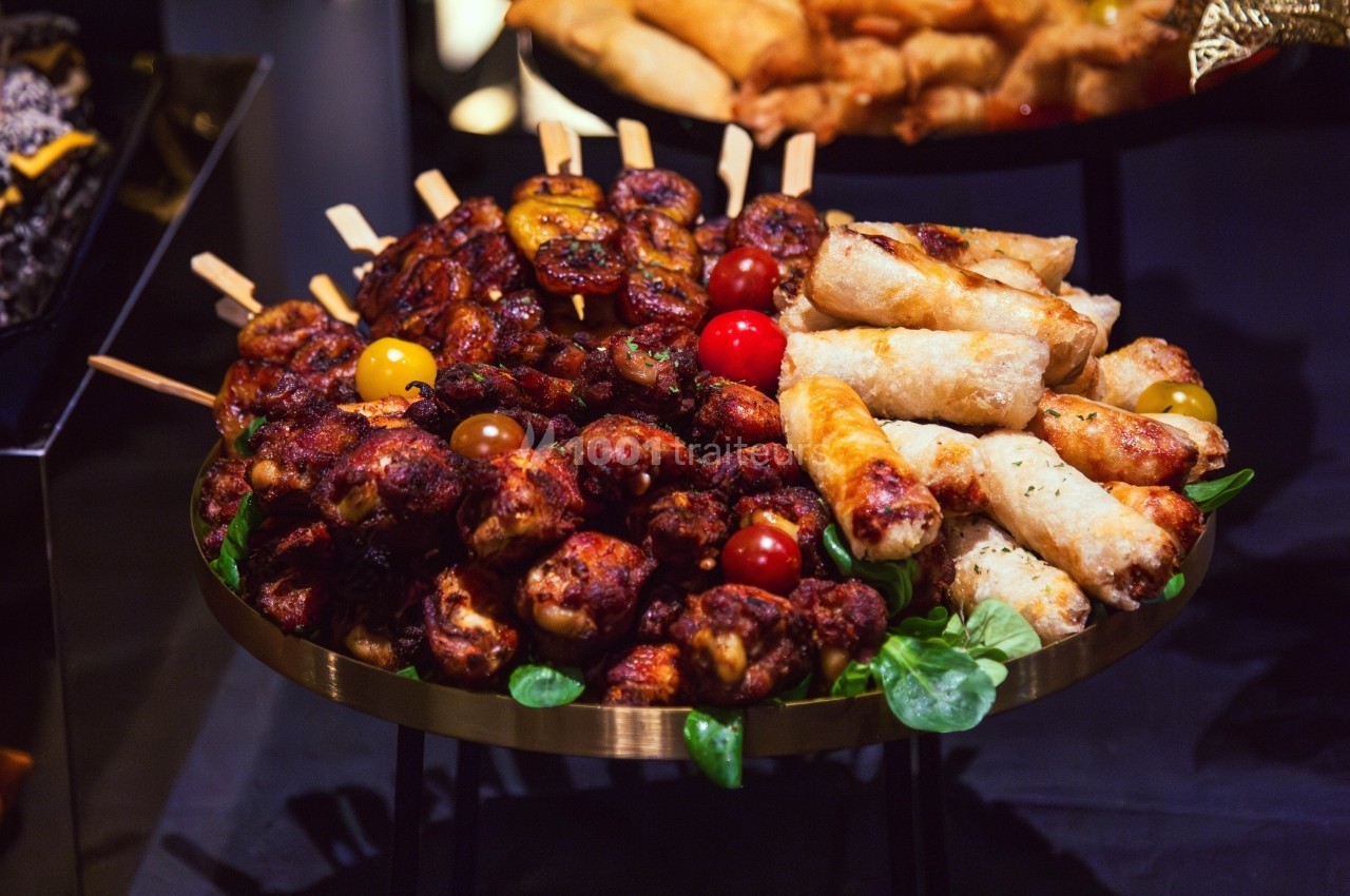 Assortiment de brochettes de viande grillée, rouleaux croustillants et légumes sur un plateau doré.
