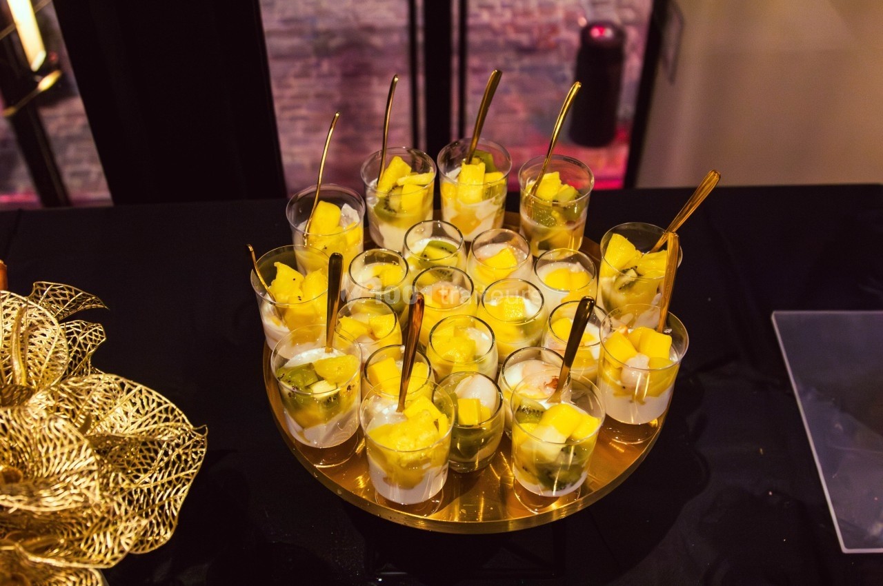Verrines de fruits frais, principalement mangue et kiwi, présentées sur un plateau doré avec des cuillères dorées.