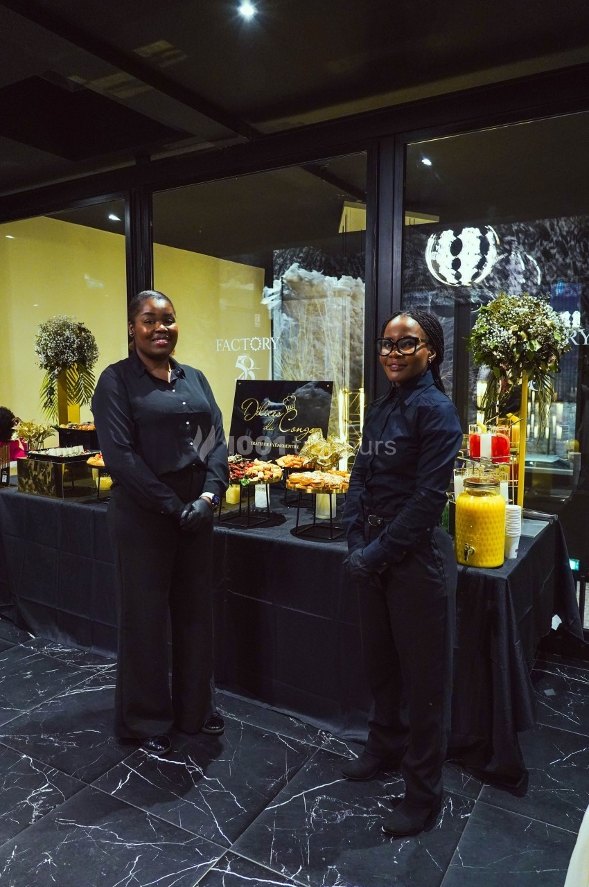 Deux personnes en tenue noire devant une table de buffet décorée avec des fleurs et des boissons.