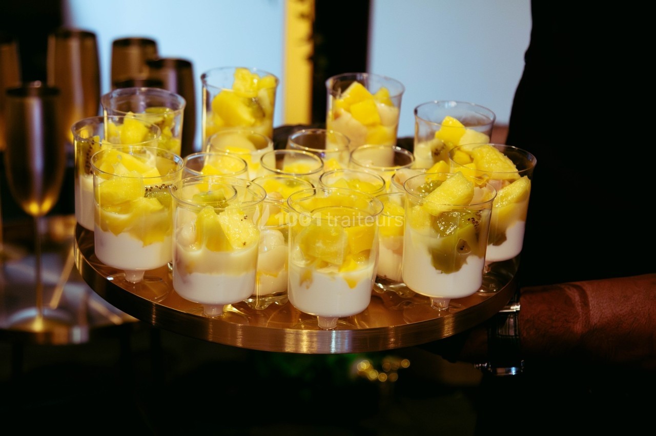 Plateau de verrines contenant du yaourt blanc et des morceaux de fruits jaunes, servi lors d'un événement.