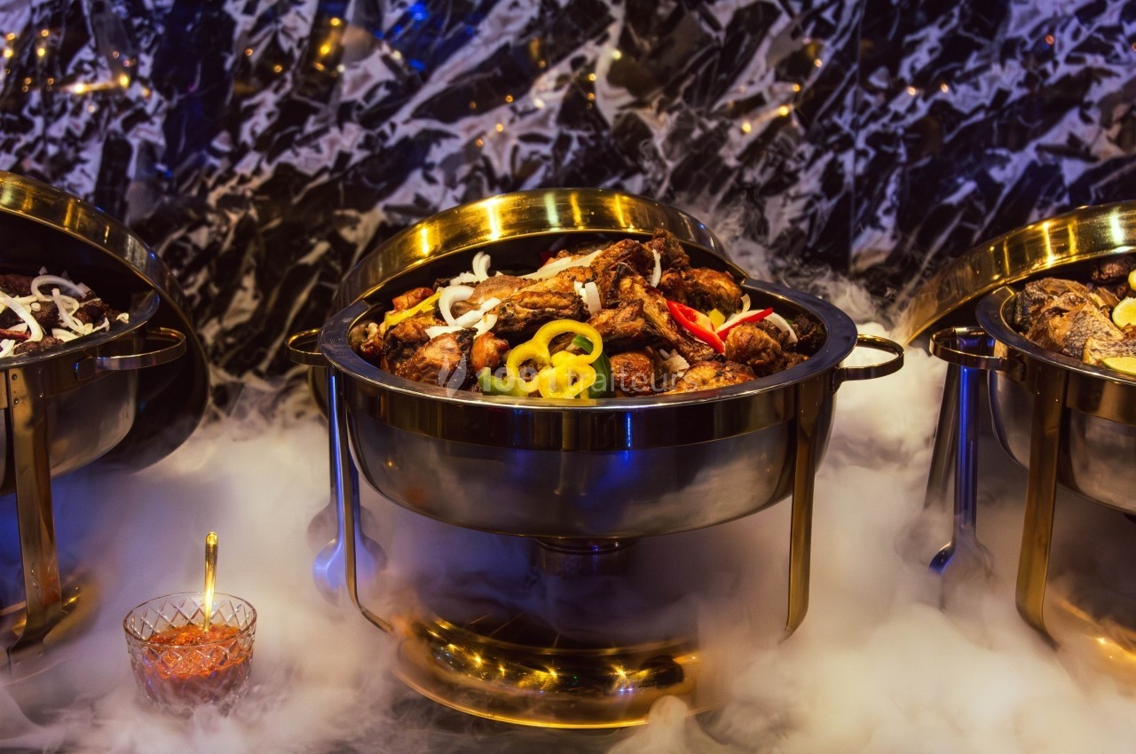 Buffet avec plats chauds présentés dans des récipients dorés, entourés de vapeur et décorés de légumes colorés.