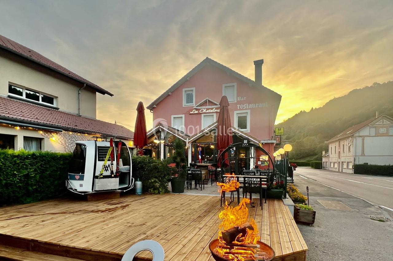 Terrasse en bois d'un restaurant avec brasero allumé, bâtiments environnants et coucher de soleil en arrière-plan.