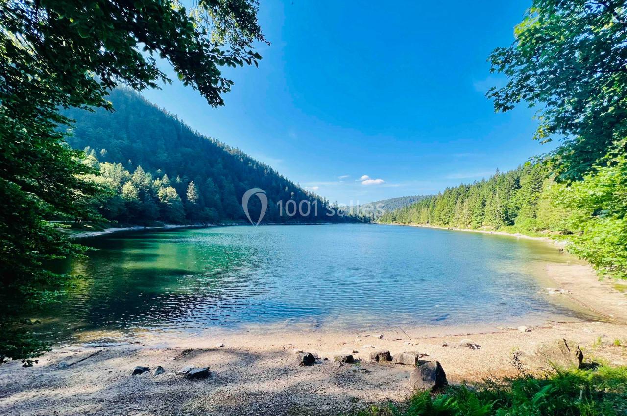 Vue d'un lac entouré de forêts et de collines sous un ciel bleu dégagé.