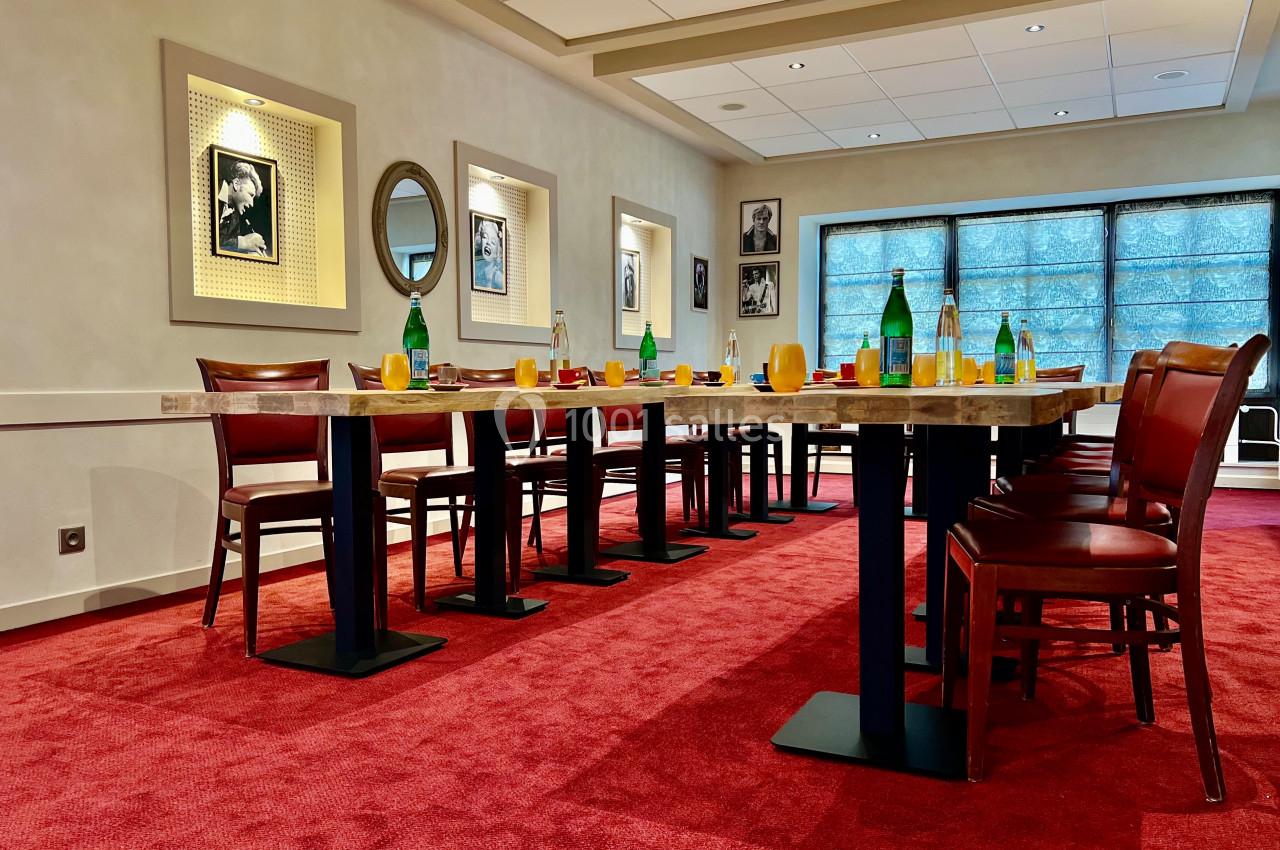 Salle de réunion avec tables alignées, chaises en bois, bouteilles d'eau et lumière naturelle filtrée par des stores.
