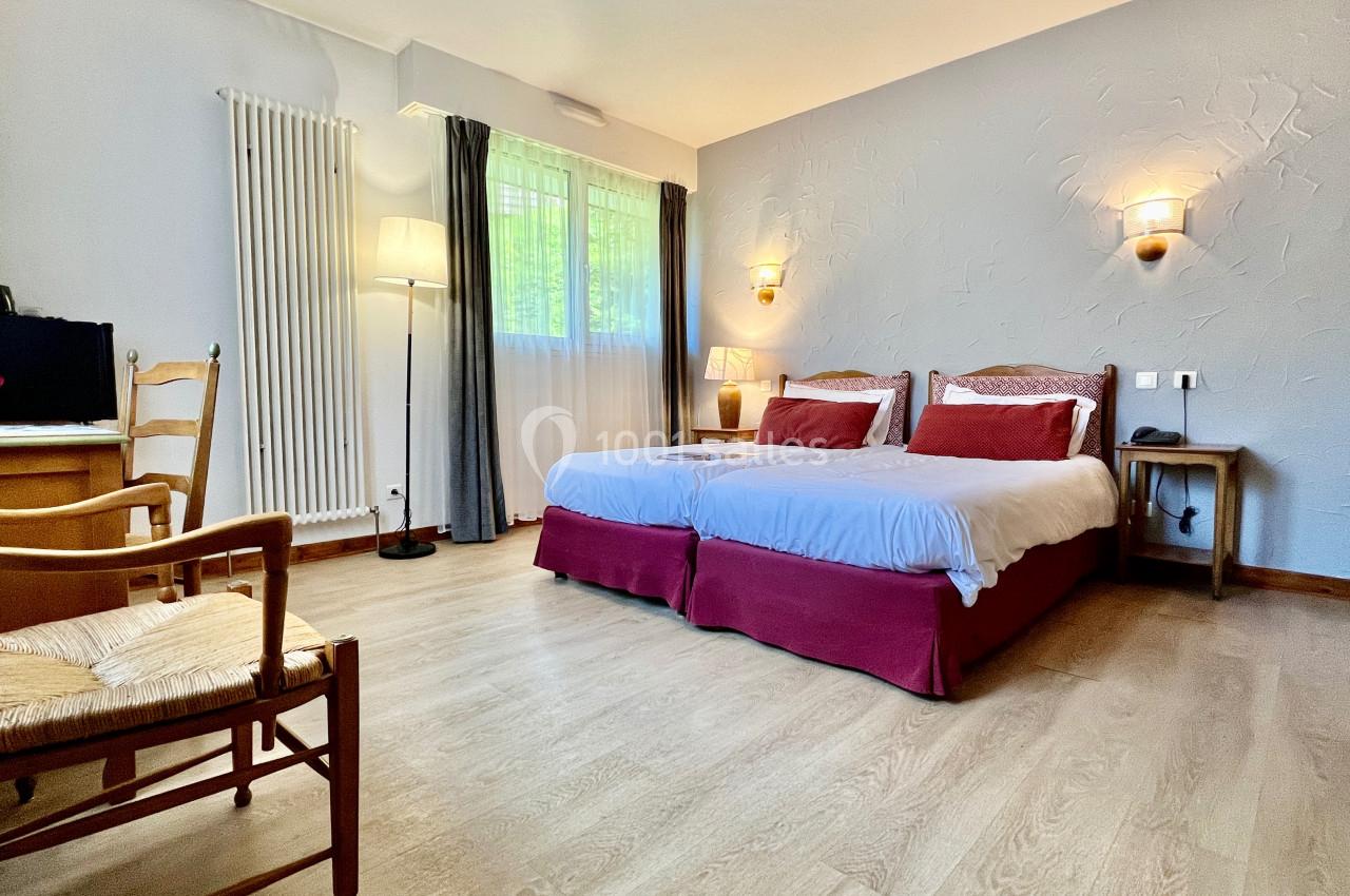 Chambre lumineuse avec deux lits simples, bureau, chaise en bois et éclairage mural, parquet au sol.