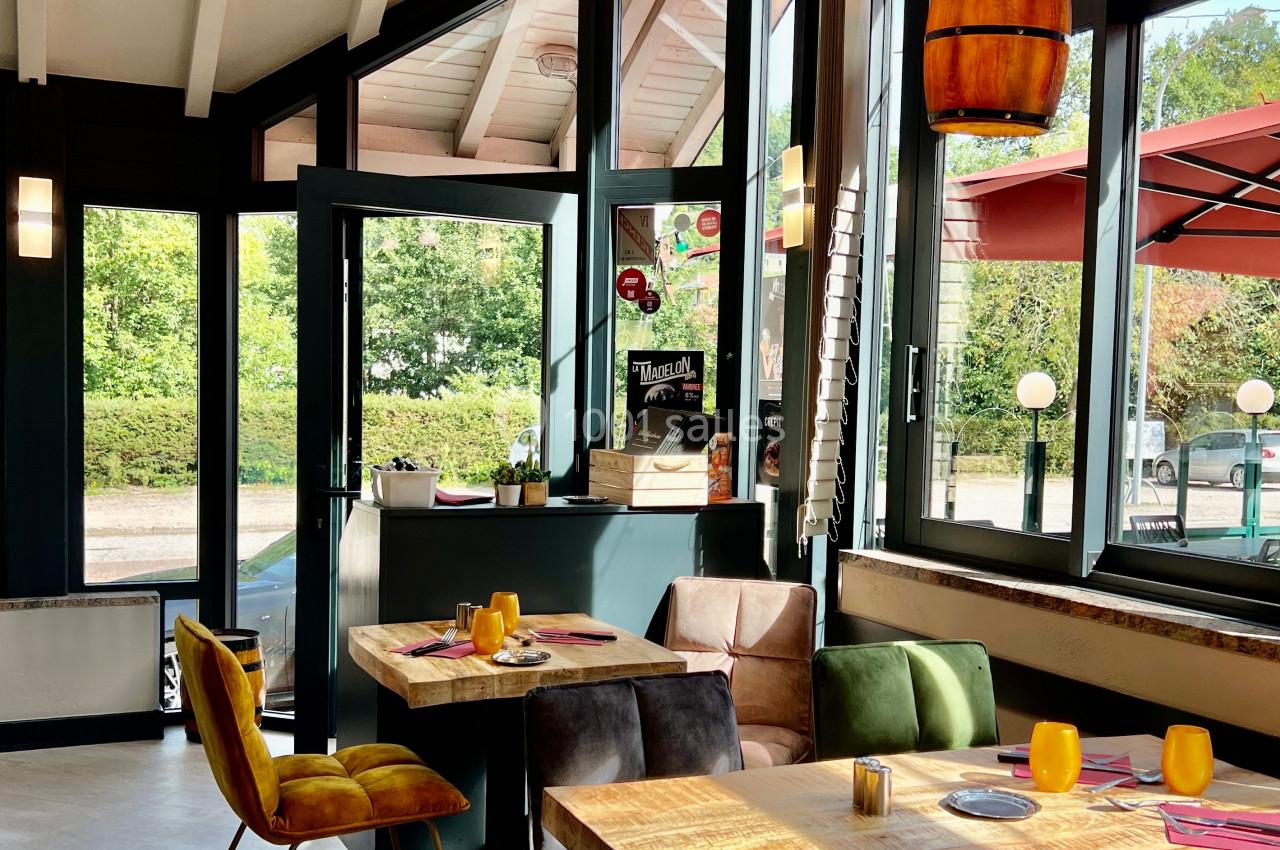 Salle de restaurant lumineuse avec grandes baies vitrées, tables en bois, chaises colorées et vue sur une terrasse…