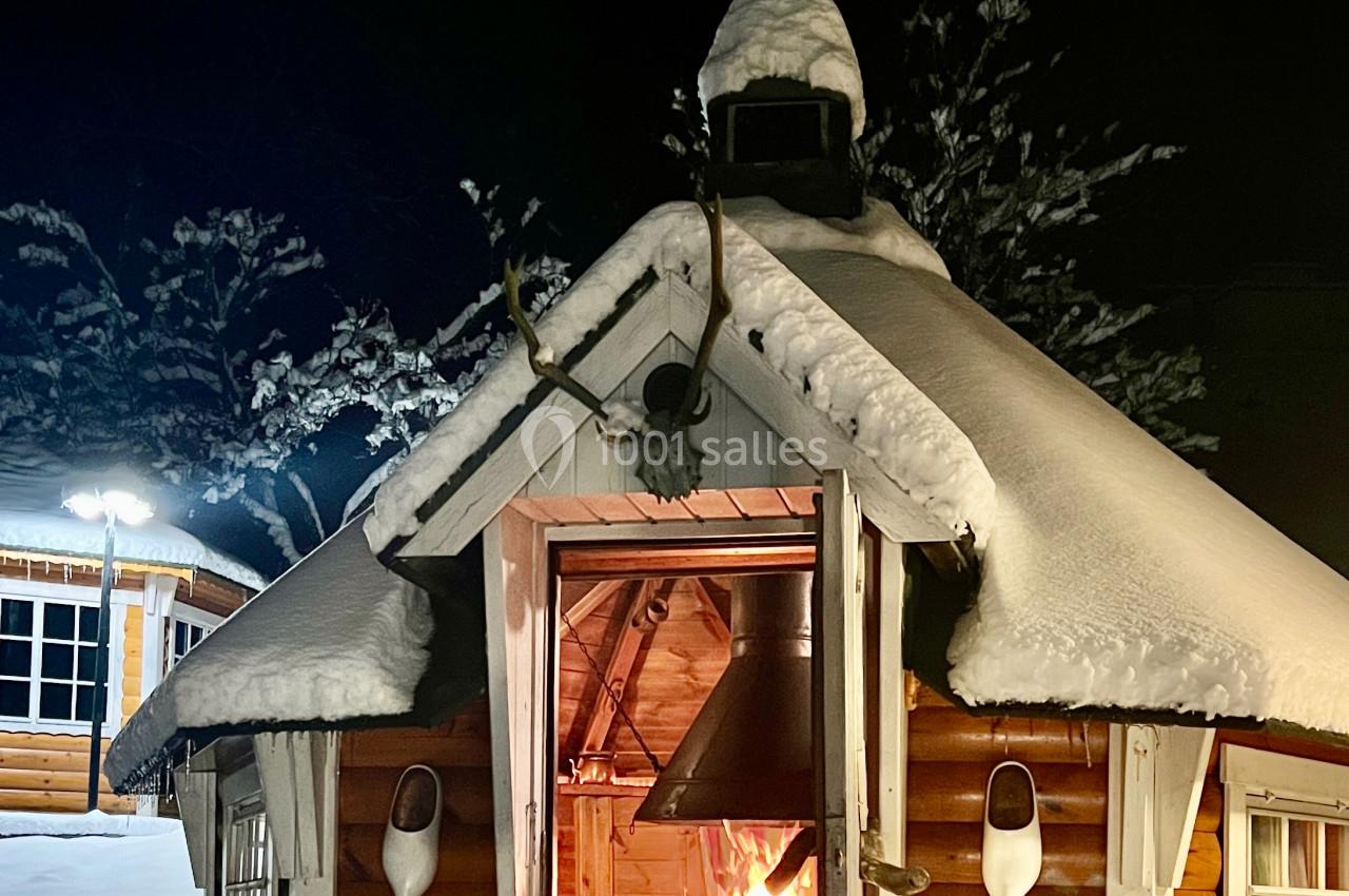Cabane en bois enneigée avec un feu allumé à l'intérieur, vue de nuit dans un paysage hivernal.