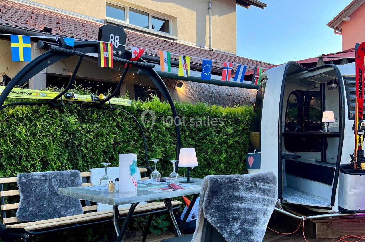 Table en extérieur avec des chaises couvertes de plaids, située près d'une cabine de télécabine décorée de drapeaux.