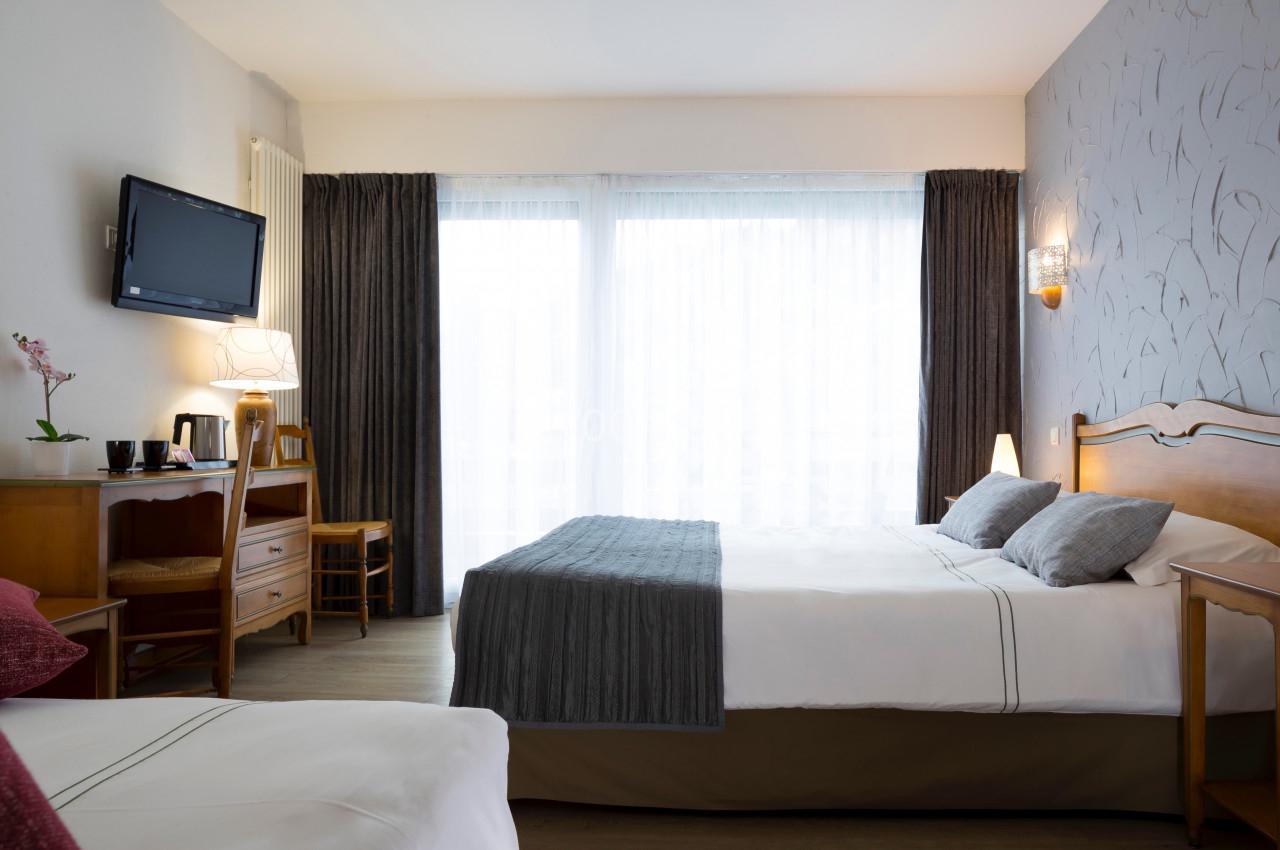 Chambre lumineuse avec lit double, bureau, télévision murale, rideaux sombres et grande fenêtre avec voilages.