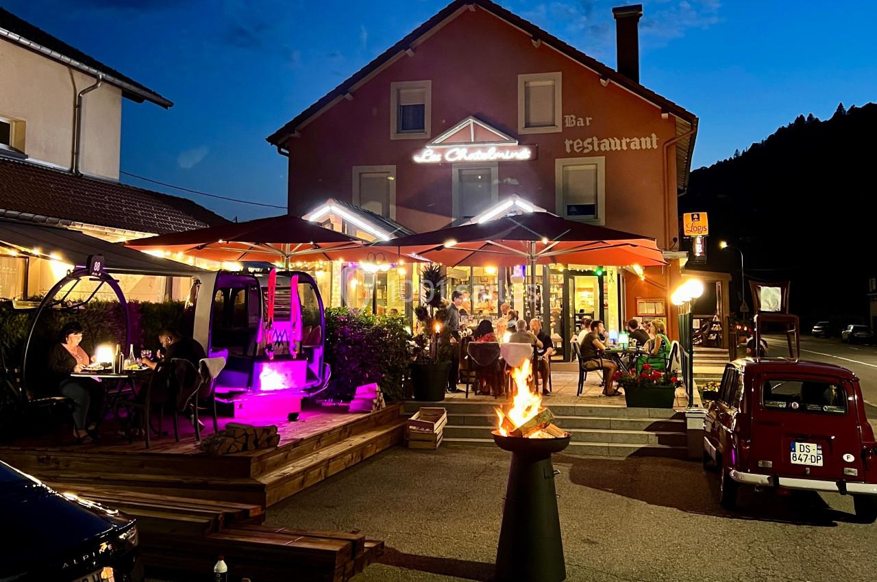 Terrasse d'un restaurant éclairée en soirée, avec des clients attablés sous des parasols et un brasero allumé au premier…