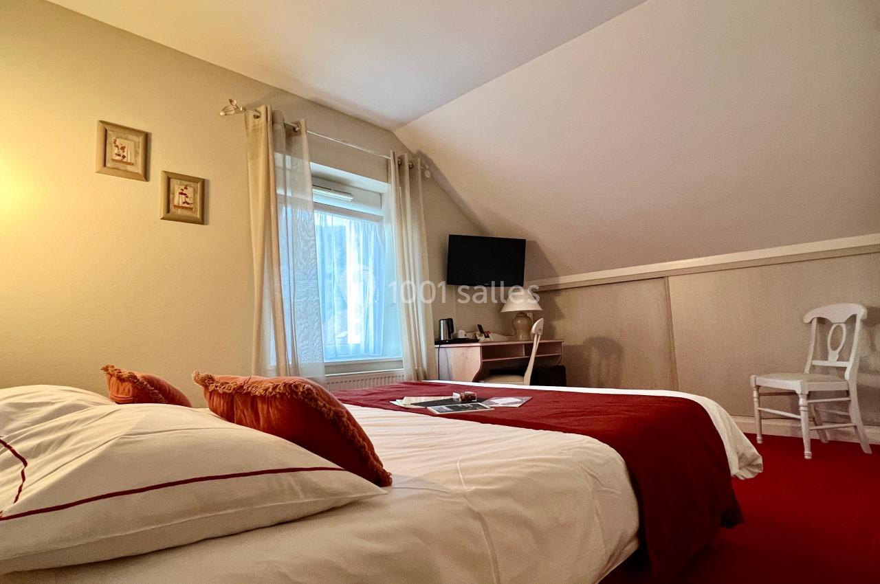 Chambre avec lit double, couvre-lit rouge, bureau avec télévision, chaise blanche et fenêtre laissant entrer la lumière…