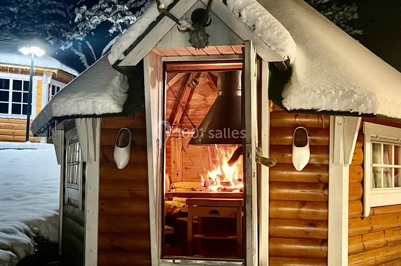 Petit chalet en bois éclairé, avec un feu allumé visible à l'intérieur, entouré de neige en soirée.