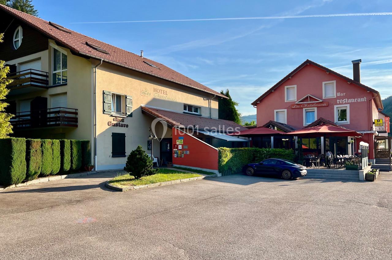 Façade d'un hôtel-restaurant avec terrasse, parking et voiture stationnée, sous un ciel dégagé.